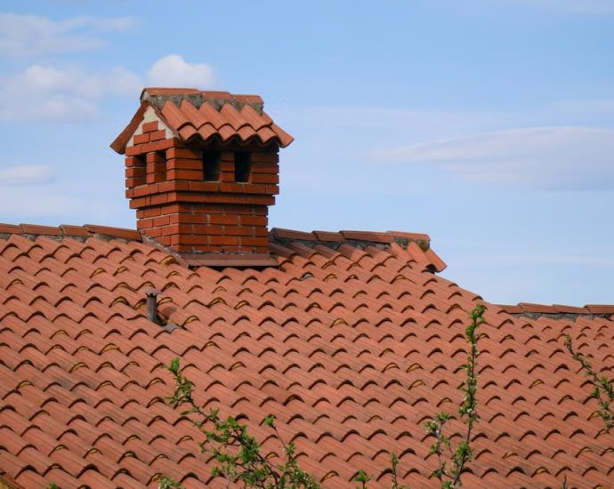 A charming red brick chimney stands atop a detailed clay-tiled roof under a clear blue sky.