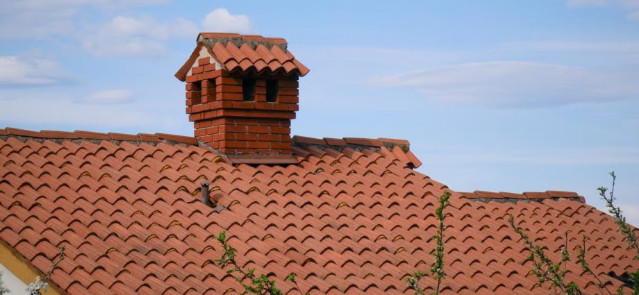 A charming red brick chimney stands atop a detailed clay-tiled roof under a clear blue sky.